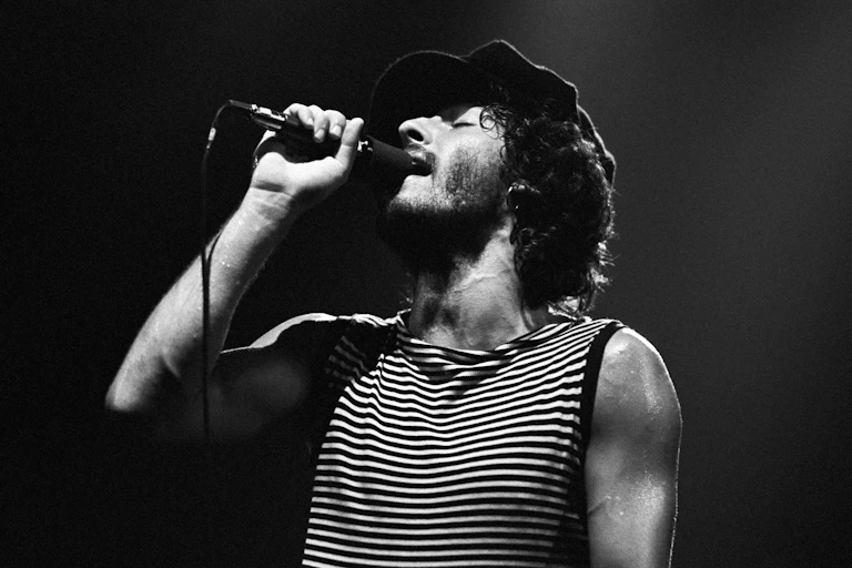 Black and white photo of young bearded man with curly hair singing into a microphone with his eyes closed.
