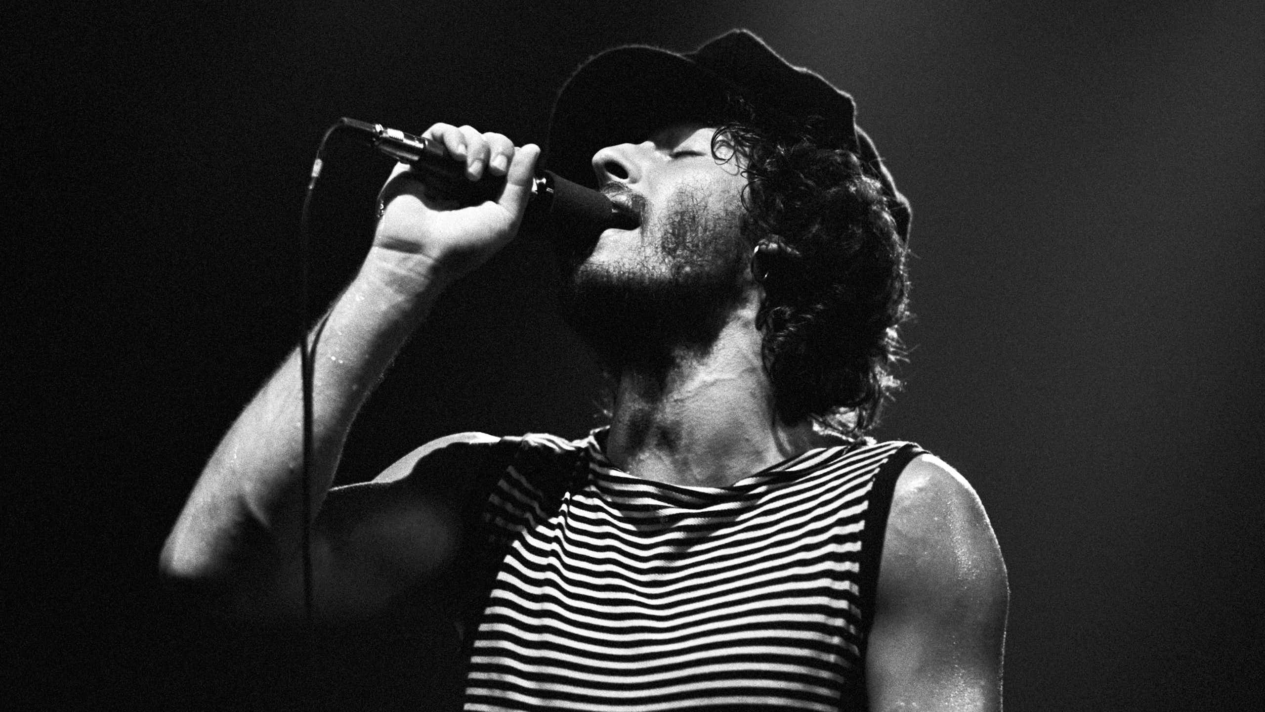 Black and white photo of young bearded man with curly hair singing into a microphone with his eyes closed.