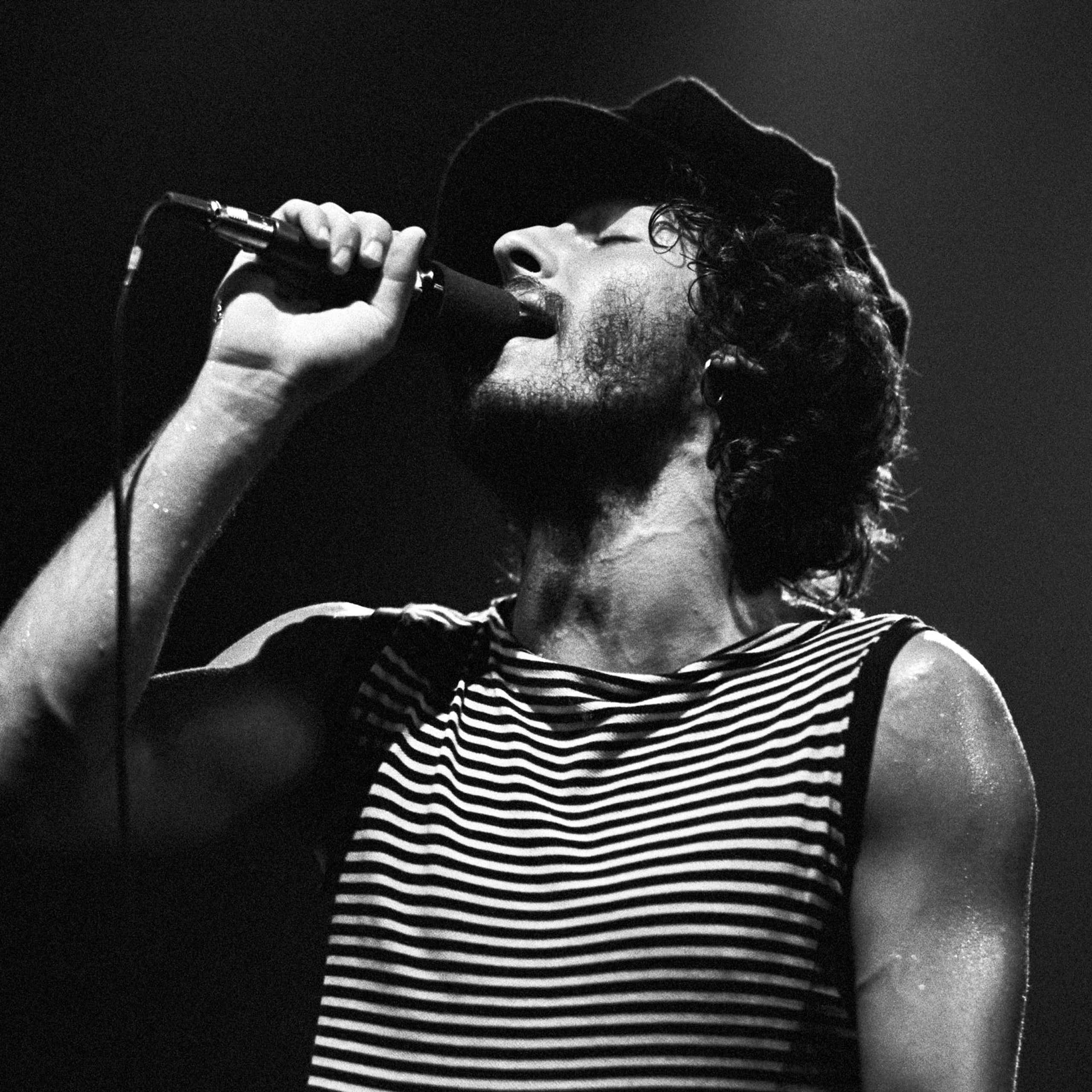 Black and white photo of young bearded man with curly hair singing into a microphone with his eyes closed.