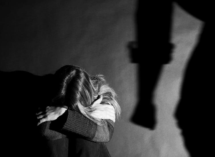 A woman on the floor, face hidden in her knees, with the looming shadow of a man’s fist beside her.