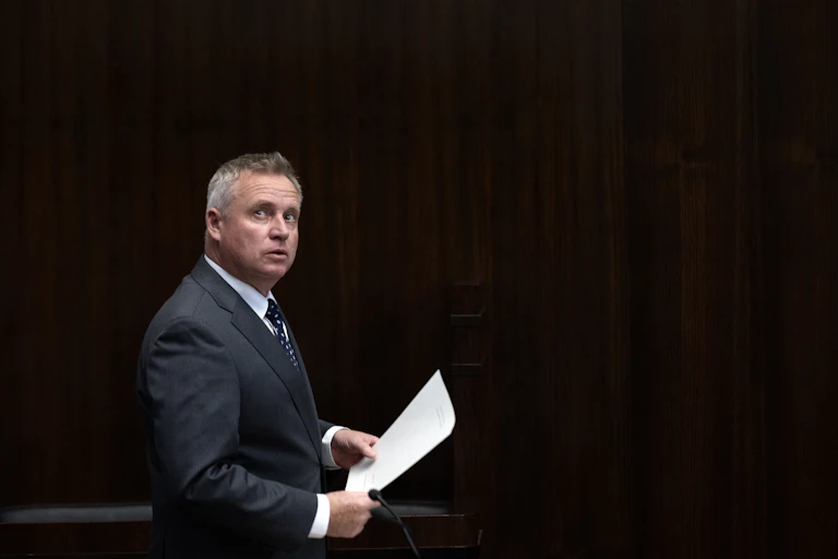 A man stands against a dark backdrop, holding papers