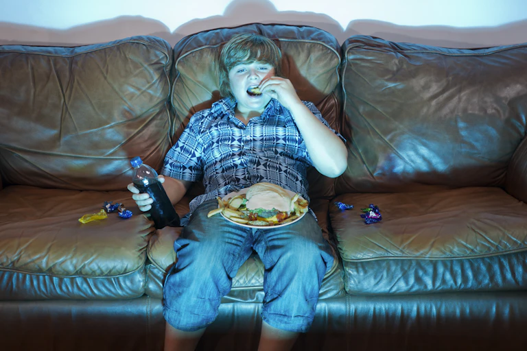 Boy eats junk food on the couch with the blue light of a screen reflected back on him.