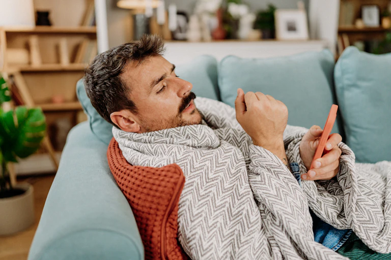 Sick man, lying on sofa, blanket around shoulder, phone in one hand