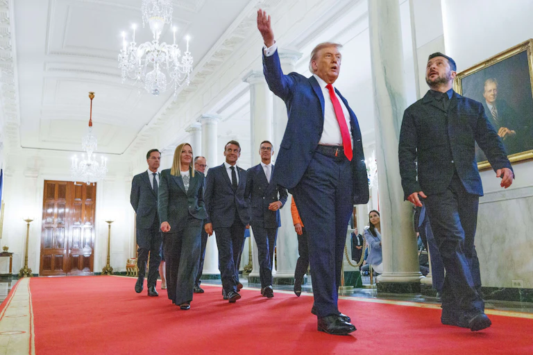 US president, Donald Trump, points to the ceiling as he walks down a White House corridor with Ukrainian president Volodymyr Zelensky. A group of aides follows.
