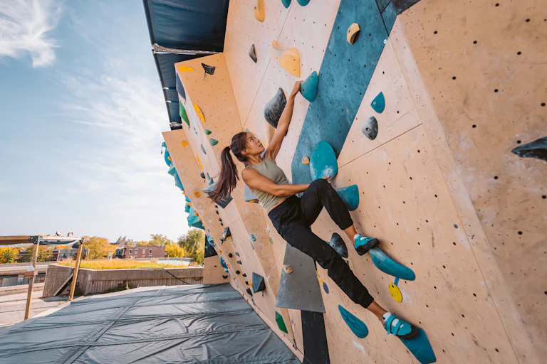 A young woman climbs a bouldering wall outdoors.