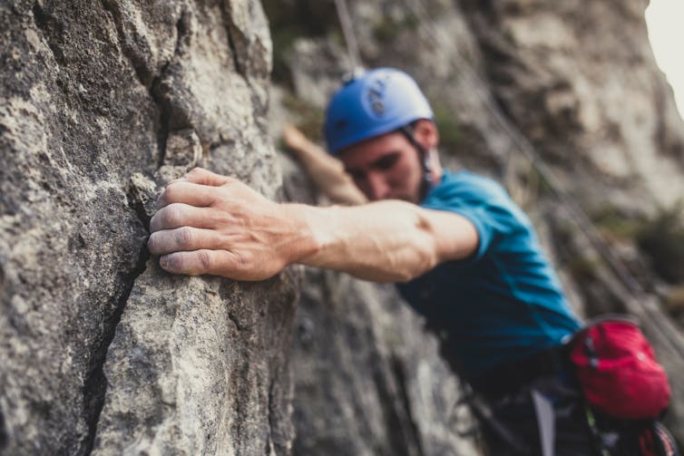 A man wearing a blue helmet tightly grips a crack in the rock face he's climbing.