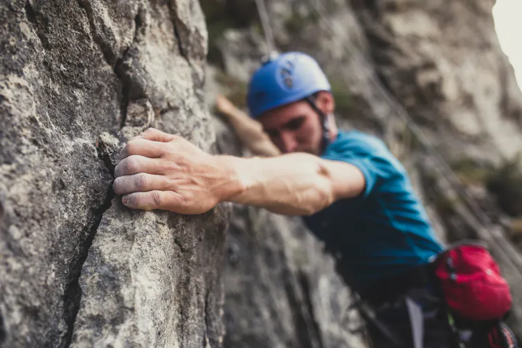 A man wearing a blue helmet tightly grips a crack in the rock face he's climbing.