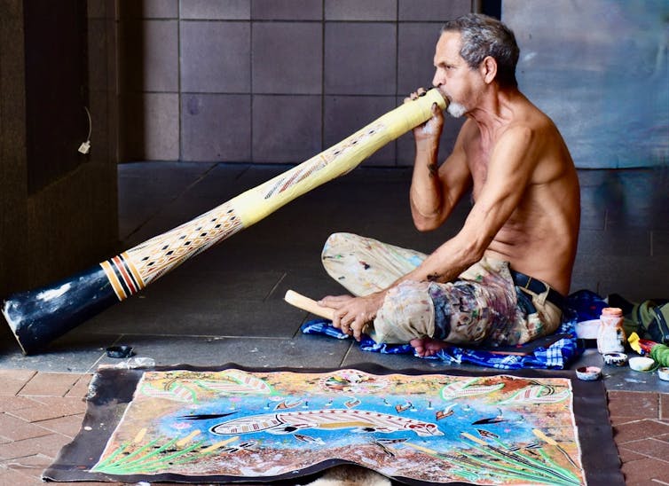 A shirtless man blows a didgeridoo.