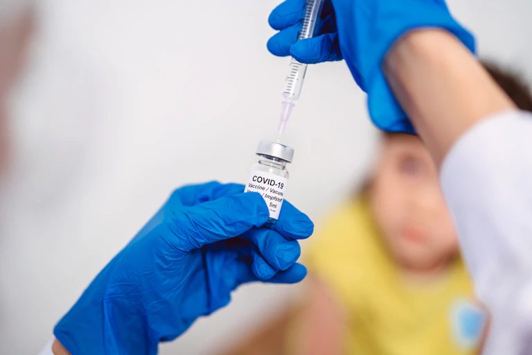 Gloved hand drawing COVID-19 vaccine out of a vial, with child in the background
