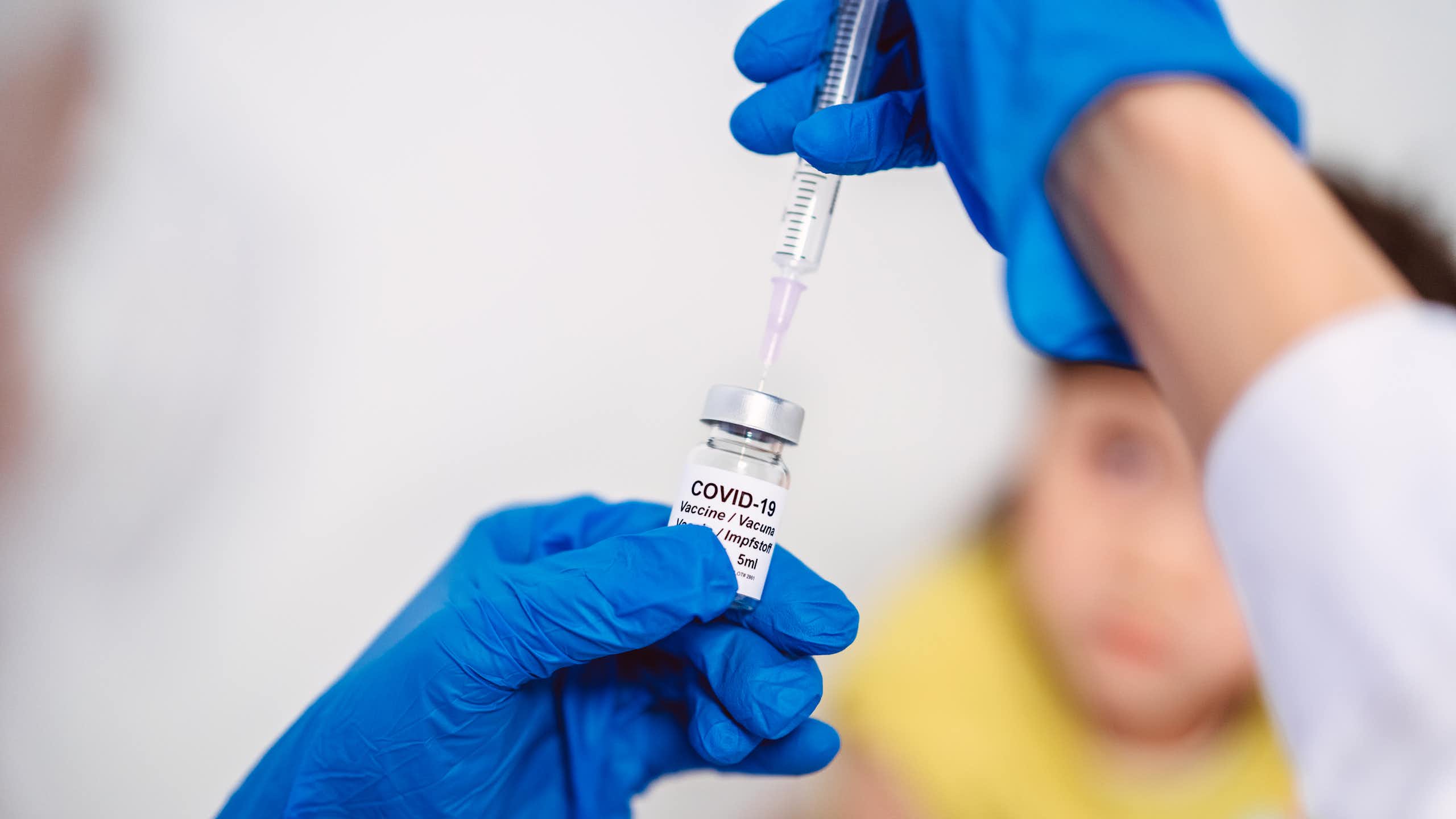 Gloved hand drawing COVID-19 vaccine out of a vial, with child in the background