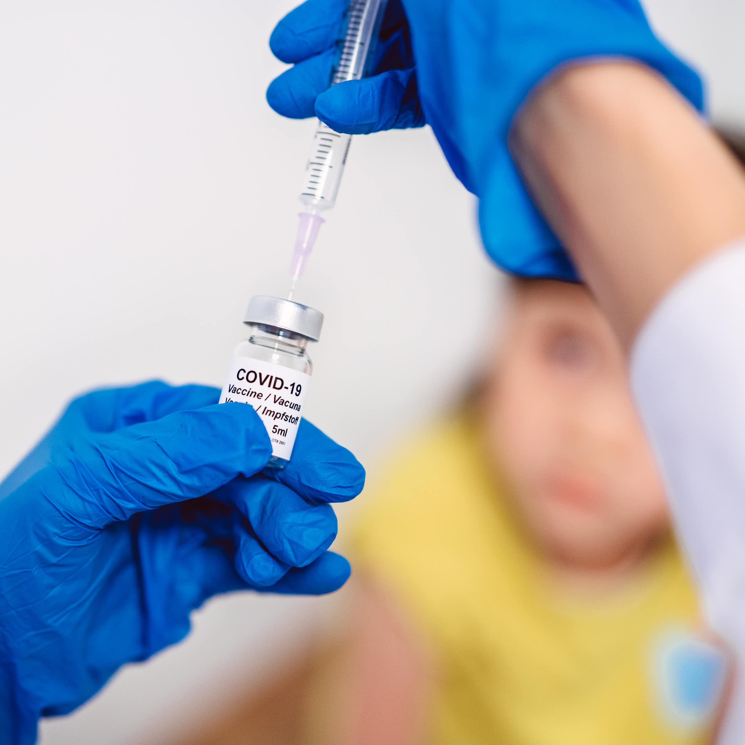 Gloved hand drawing COVID-19 vaccine out of a vial, with child in the background
