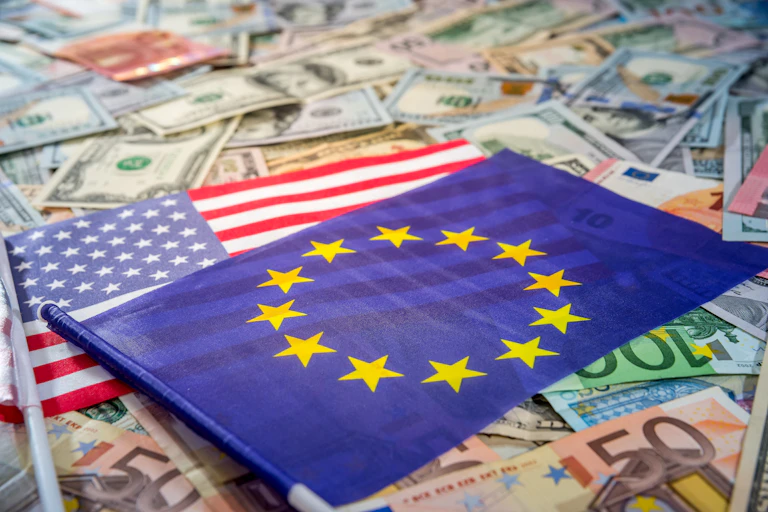 The EU flag is shown on top of an American flag, which is spread atop a bed of euros and dollar bills.