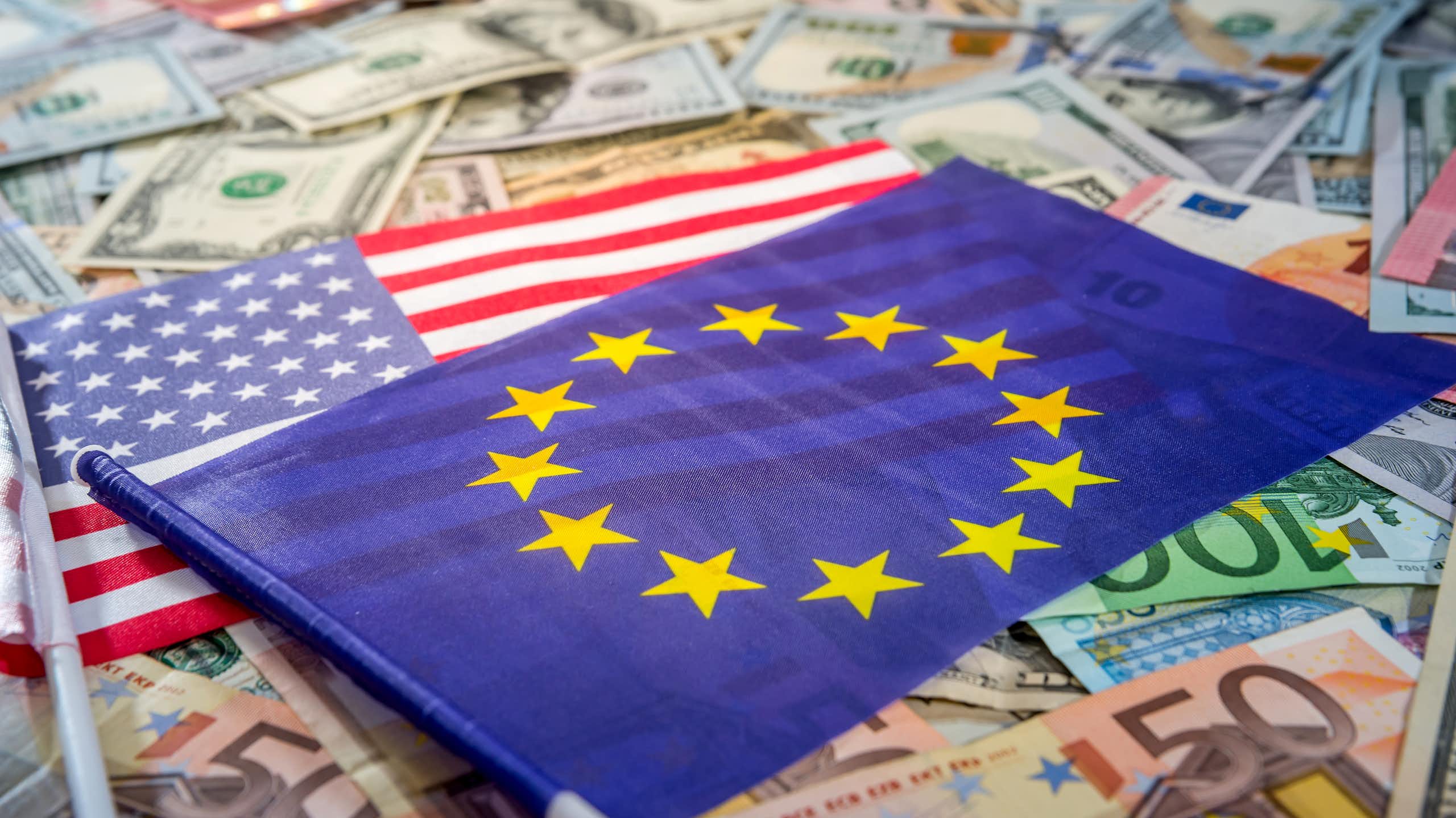 The EU flag is shown on top of an American flag, which is spread atop a bed of euros and dollar bills.