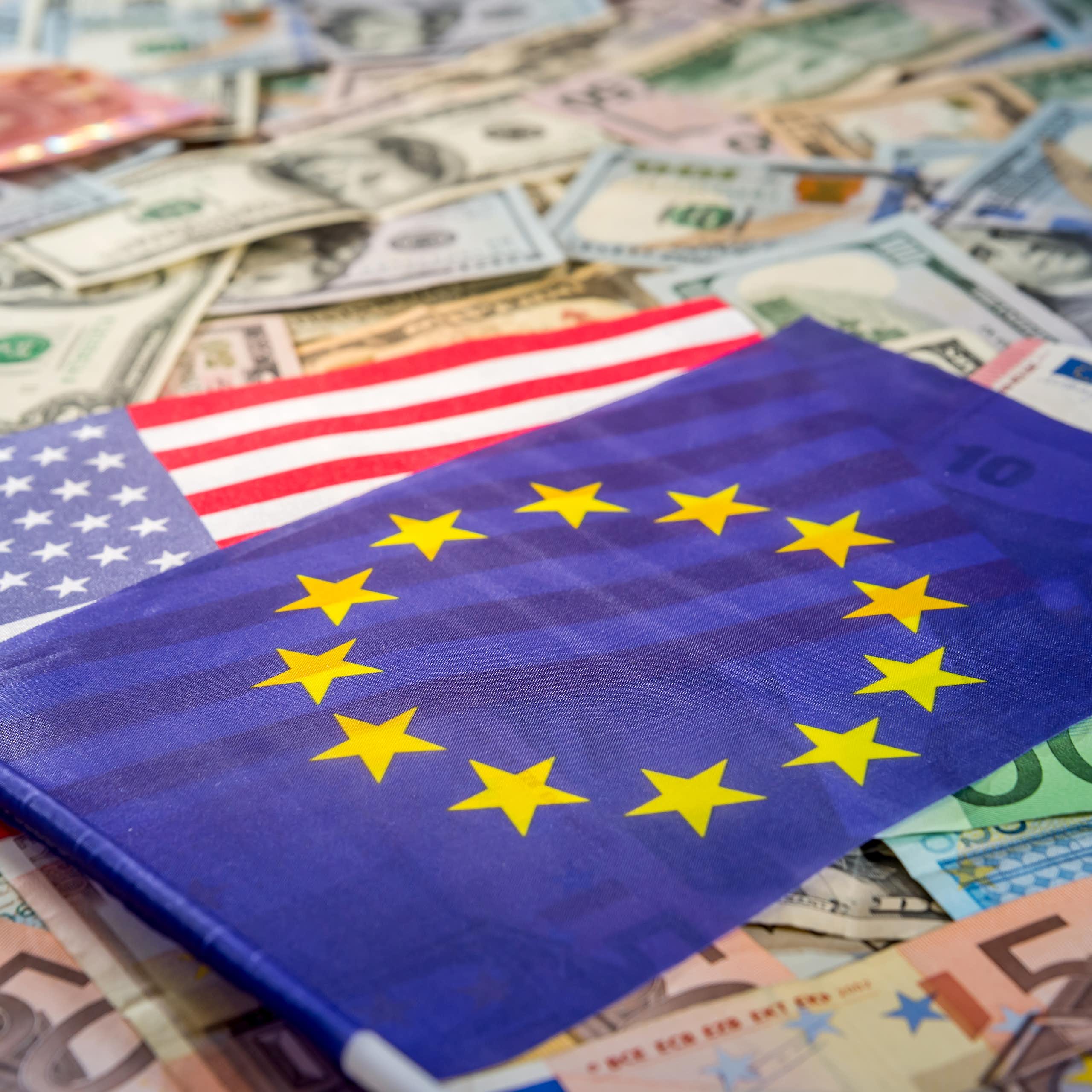 The EU flag is shown on top of an American flag, which is spread atop a bed of euros and dollar bills.