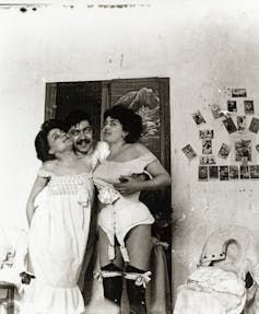 A black and white photo of a man holding a woman on either side of him, in their old fashioned underwear.