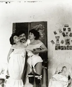 A black and white photo of a man holding a woman on either side of him, in their old fashioned underwear.