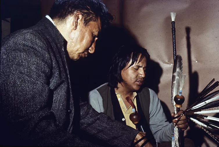 Two men, holding feathers and a small attached pot, praying with their eyes closed.