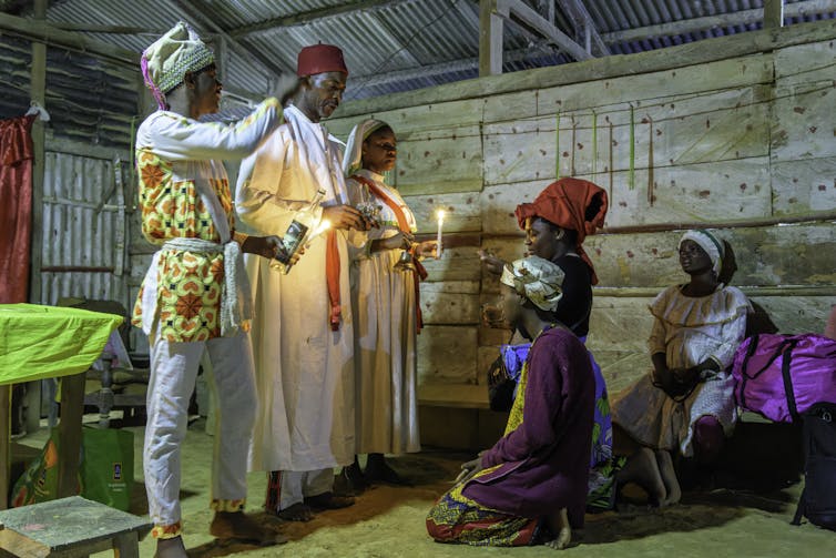 Several people wearing headdresses and long robes appear to be blessing others, while some are kneeling before them.