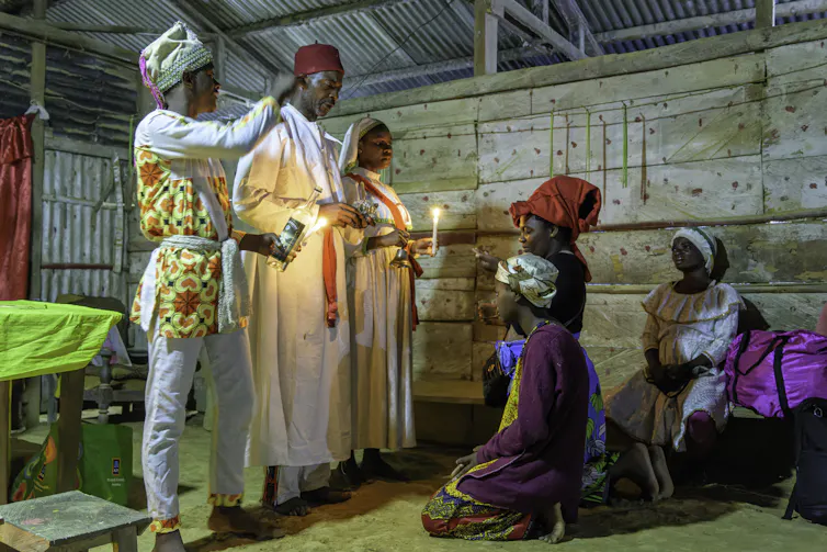 Several people wearing headdresses and long robes appear to be blessing others, while some are kneeling before them.
