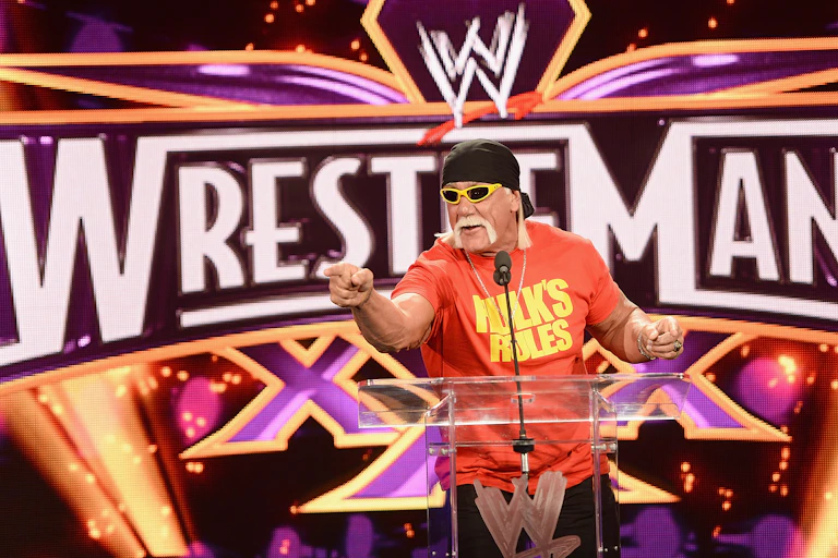 Man with a big mustache points at the camera with a backdrop that says 'Wrestlemania.'