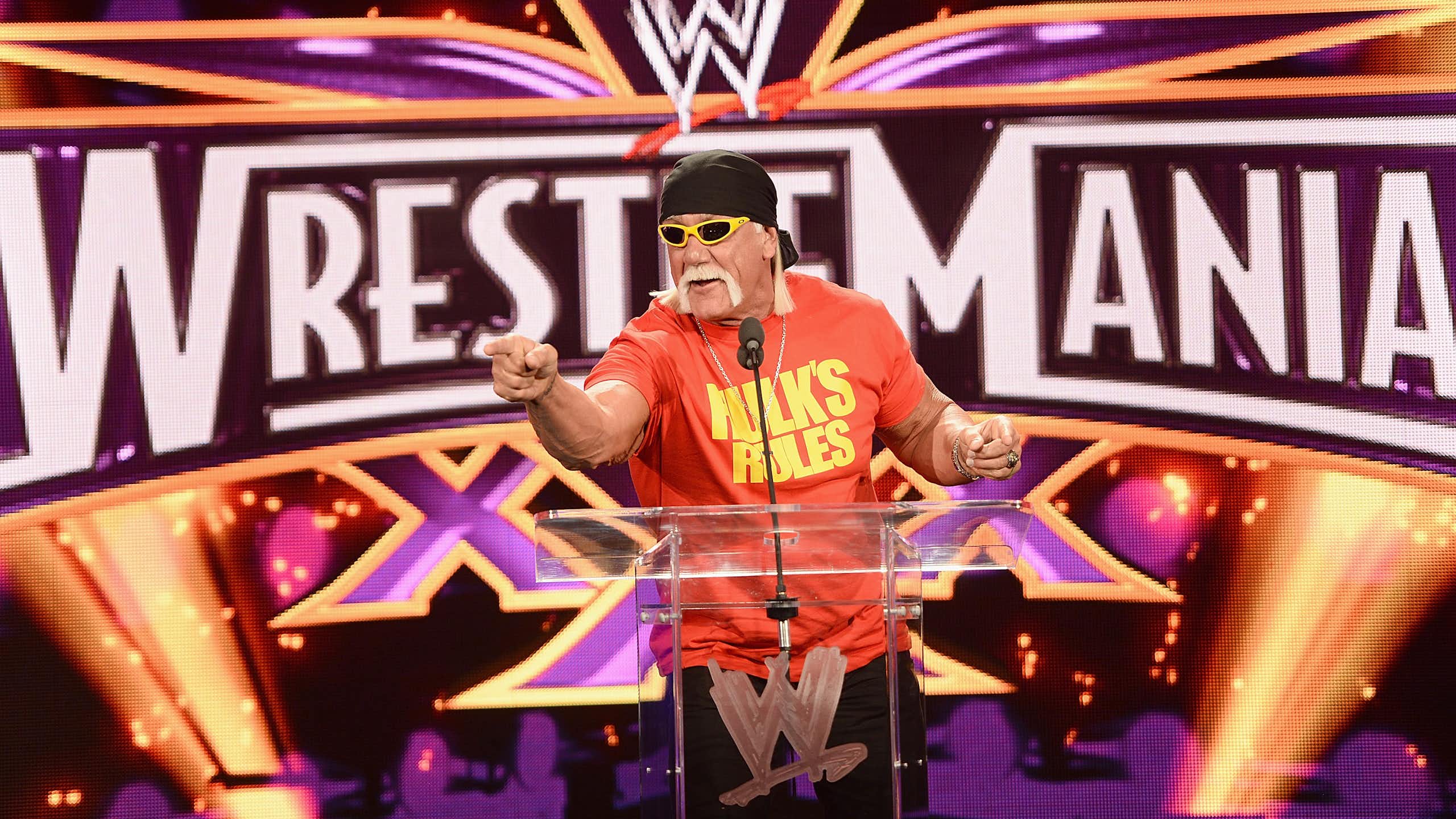 Man with a big mustache points at the camera with a backdrop that says 'Wrestlemania.'