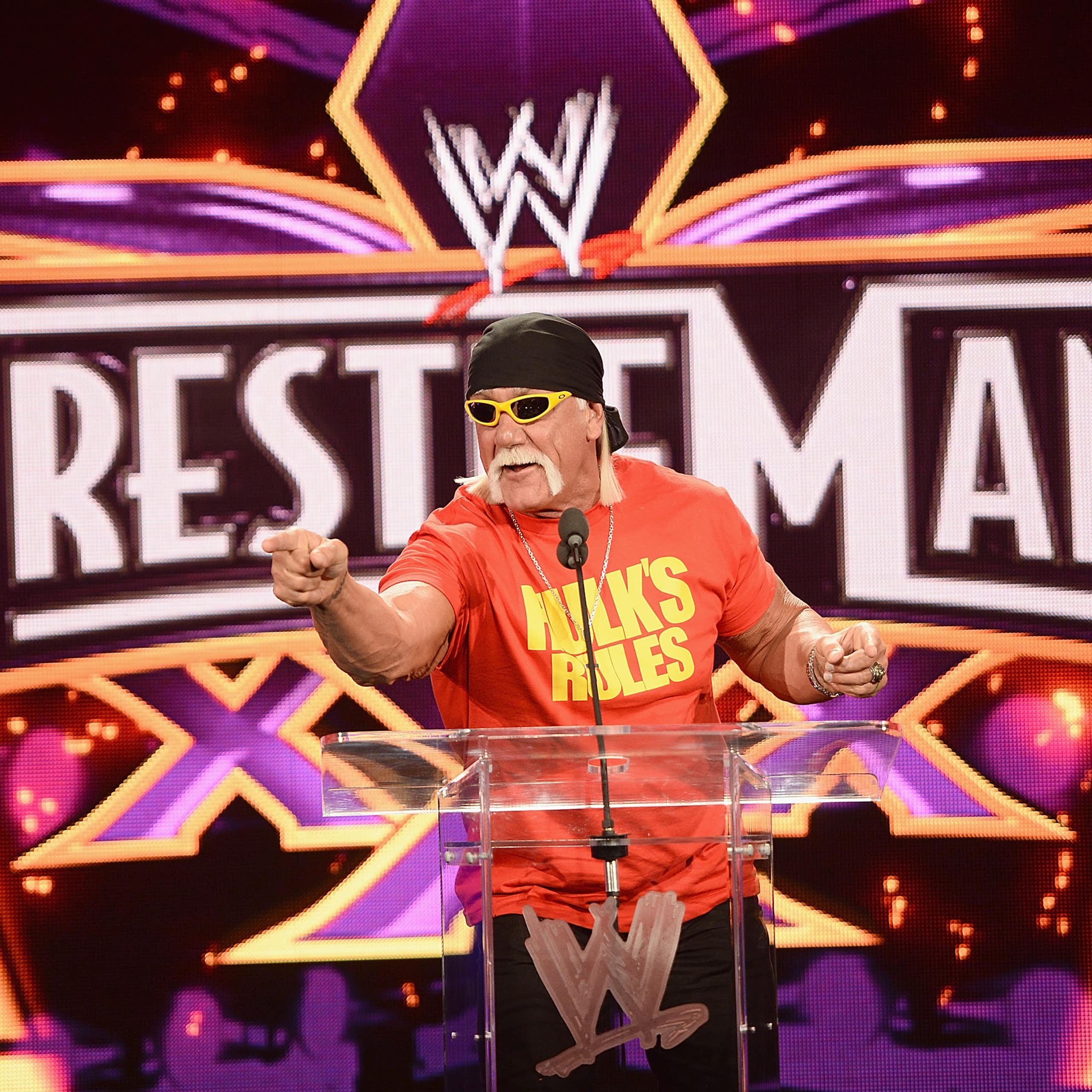 Man with a big mustache points at the camera with a backdrop that says 'Wrestlemania.'