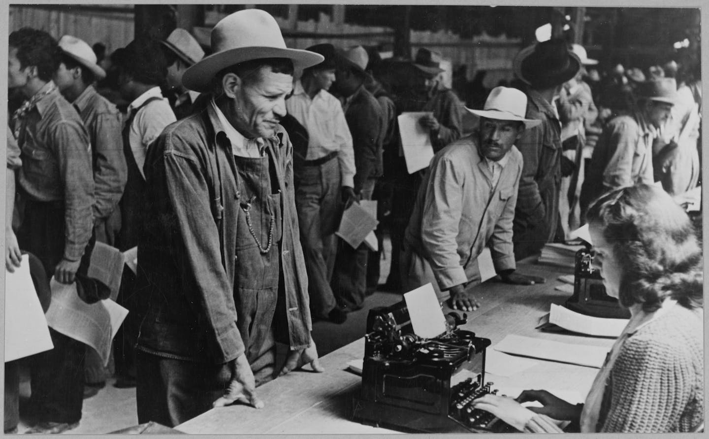 A black and white photos shows several men standing in front of a table as a woman sits on the other side of the table.