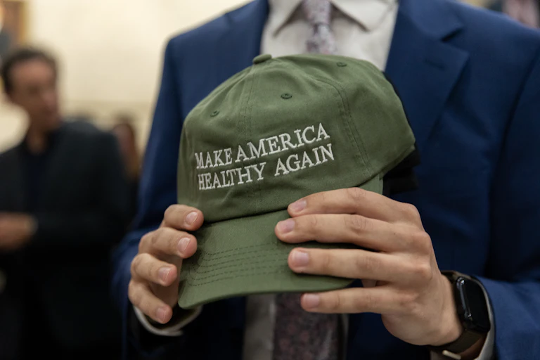 Headless man in suit jacket holding an army green baseball cap that says "MAKE AMERICA HEALTHY AGAIN"