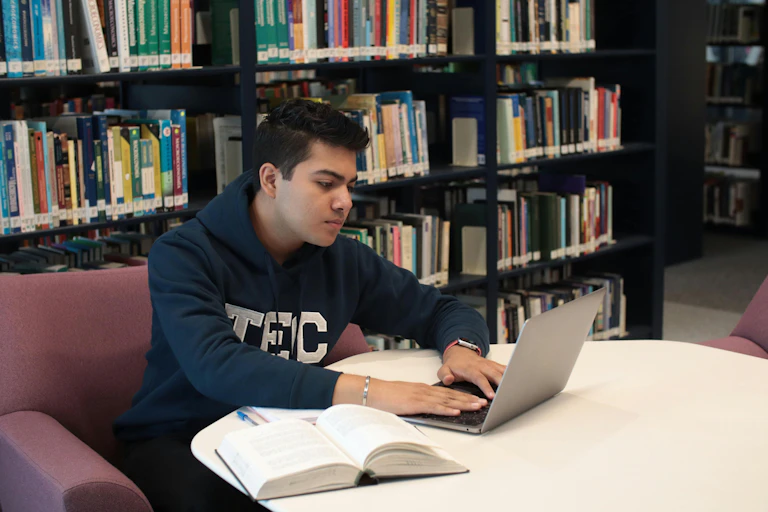 A student works in a library, typing on a laptop with a thick book open next to him.
