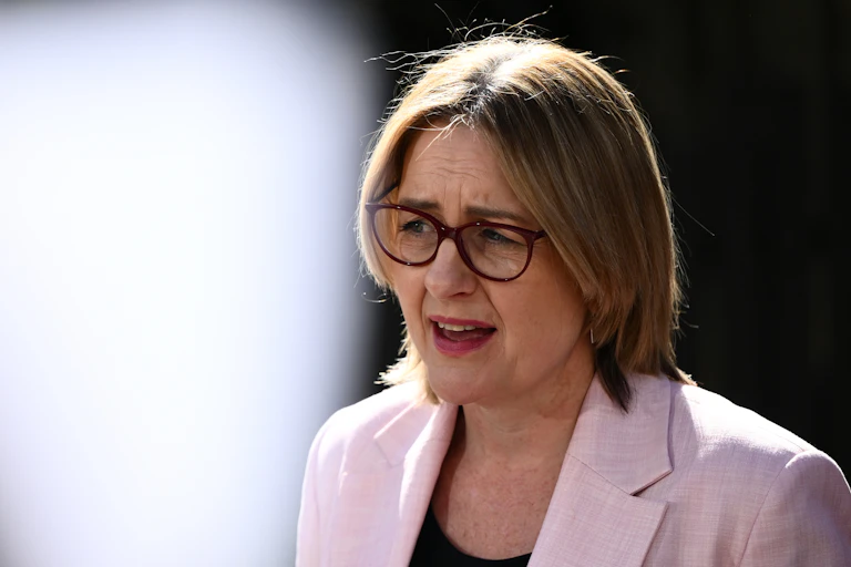 A woman in glasses and a pale pink blazer addresses the media