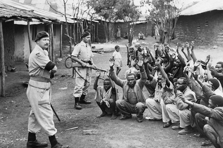 Black and white photo of armed men in uniform standing over a group of people who are squatting on the ground with their arms held above their heads