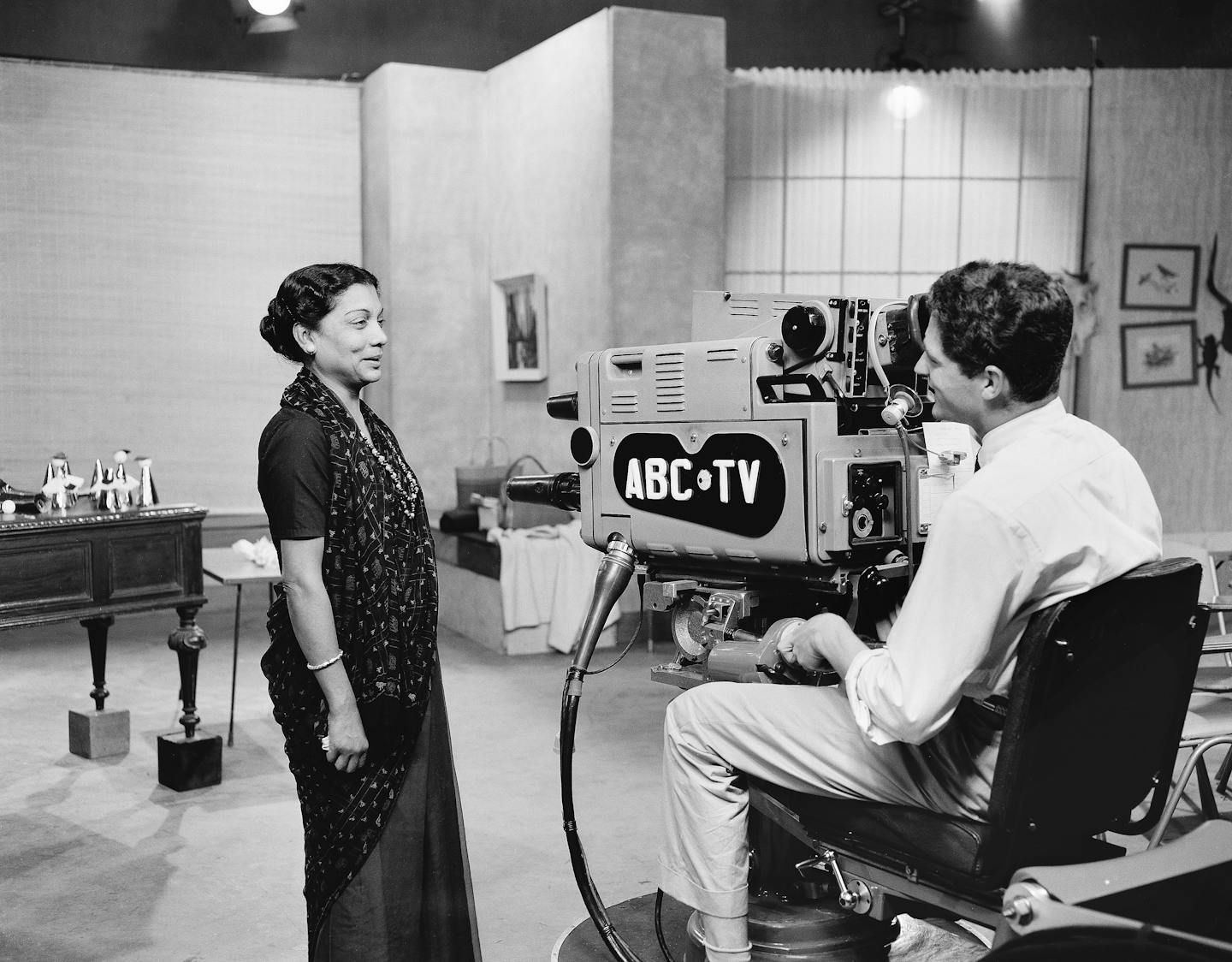 A figure stands in a television studio, talking with a camera operator who is sitting with a television camera.