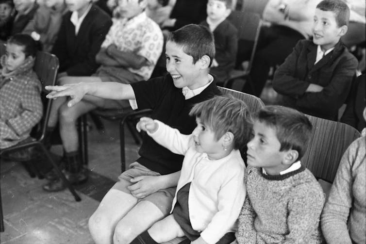 A group of children seated in a room. Two children point at a television screen out of frame.