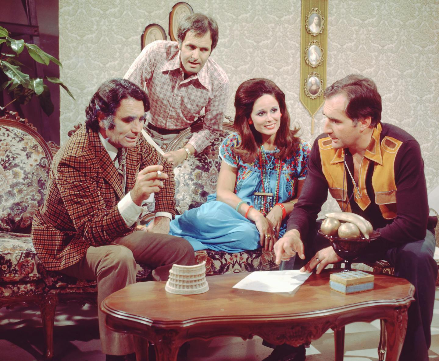 Four figures surround a desk. Each wears colourful clothes from the 1970s.