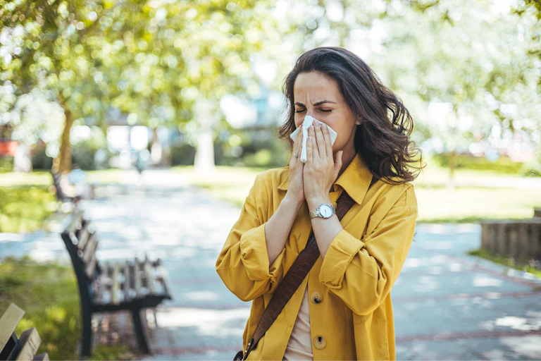 A woman stands in an outdoor park and blow her nose into a tissue.
