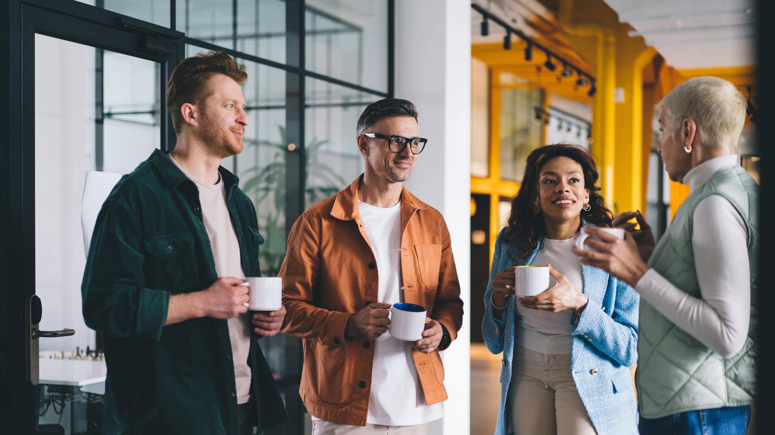 Four adults are talking while holding mugs or cups in an office setting.