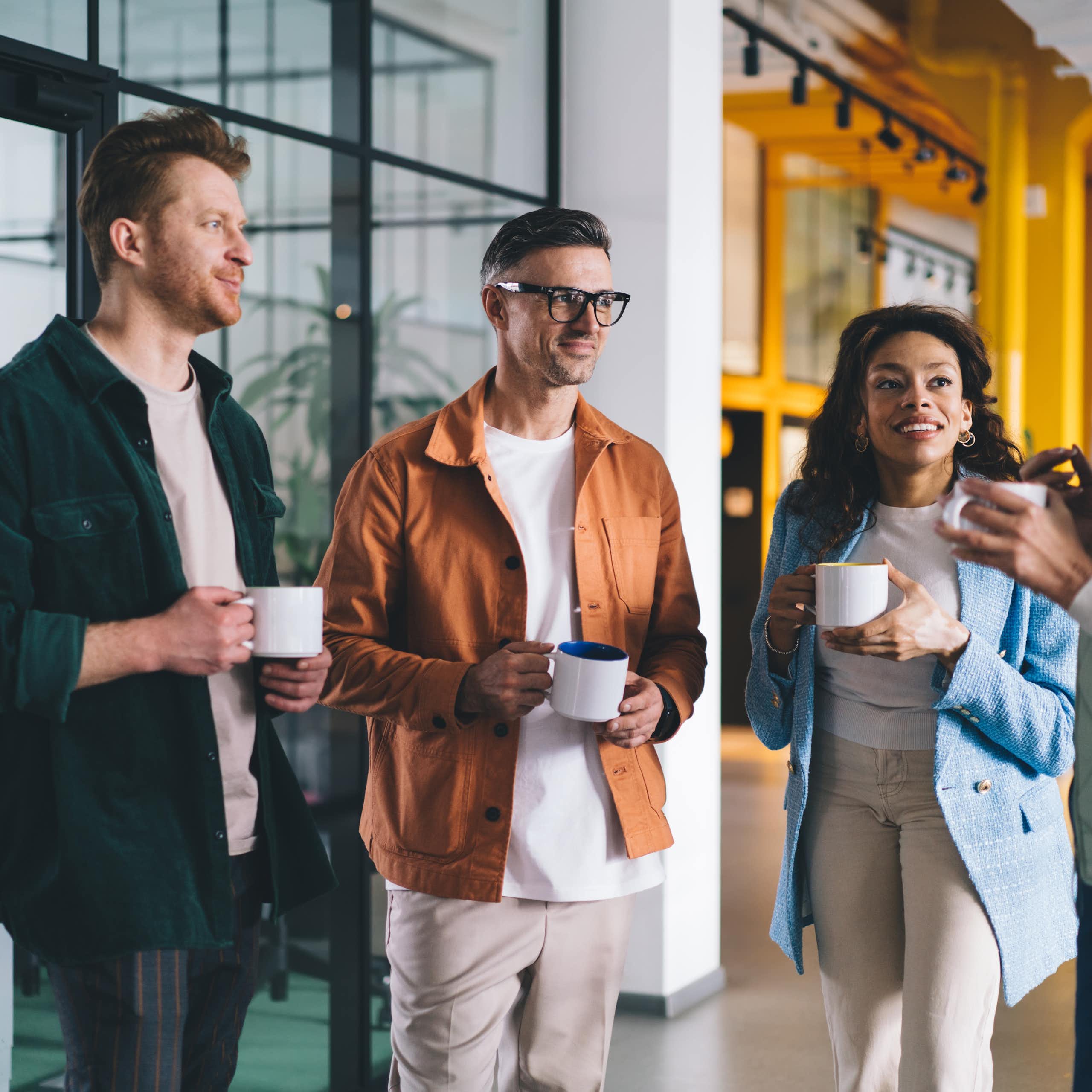 Four adults are talking while holding mugs or cups in an office setting.