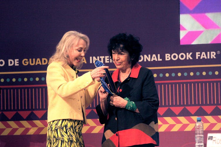 A woman with blonde hair presents a medal on a stage to an older woman with short, black, curly hair.