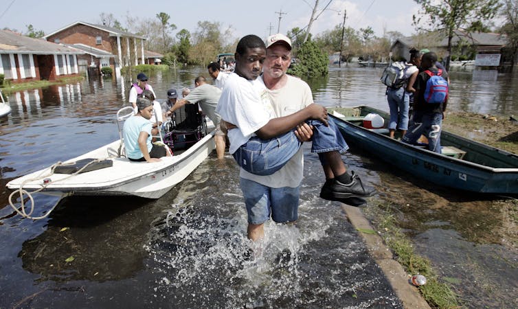 A man carries a smaller man from a boat to dry land while people wait in the boat behind him on a flooded city street.