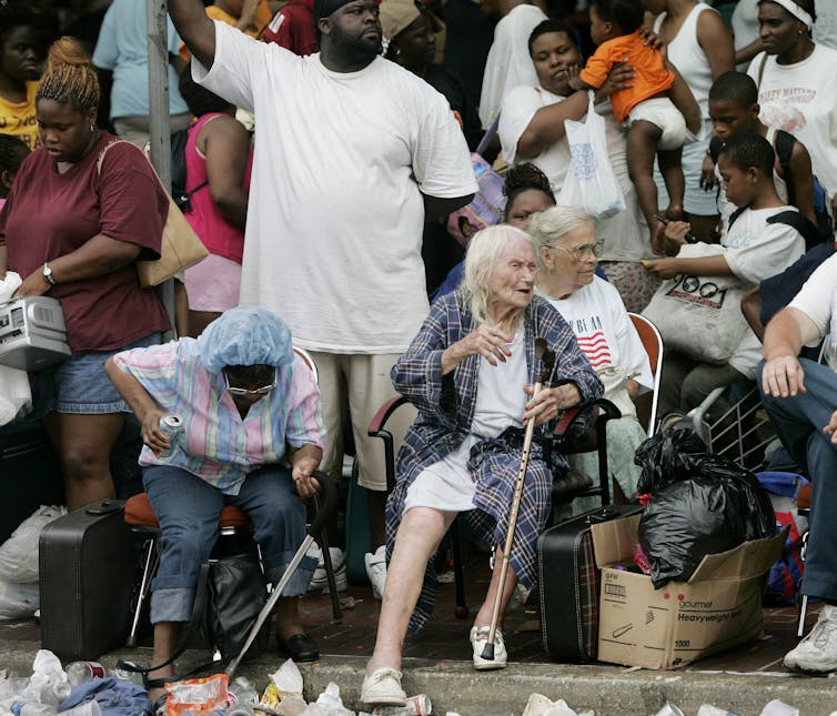 Three older women in portable chairs look for arriving transportation. Many more people crowd the curb around them.