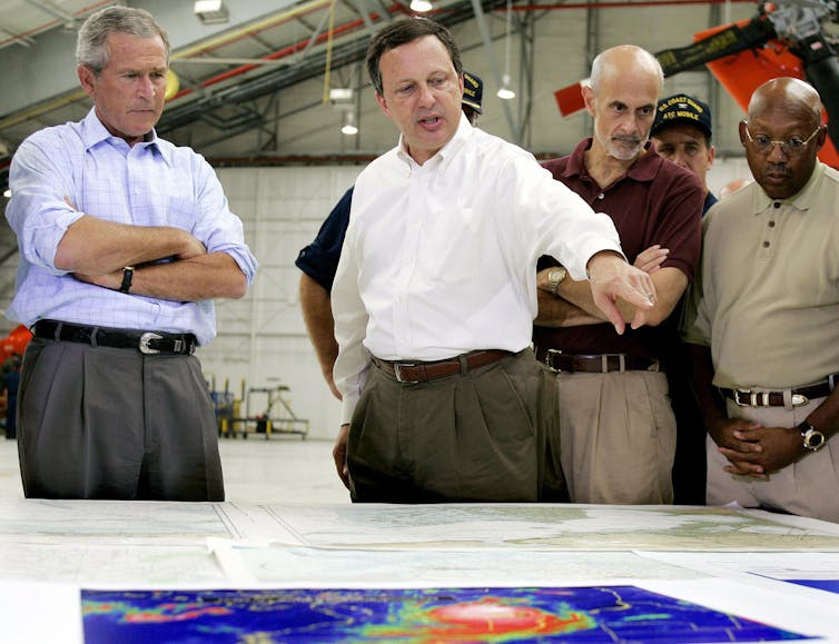 A man in party rolled up shirt sleeves points to a map while President George W. Bush stands listening nearby with his arms crossed.