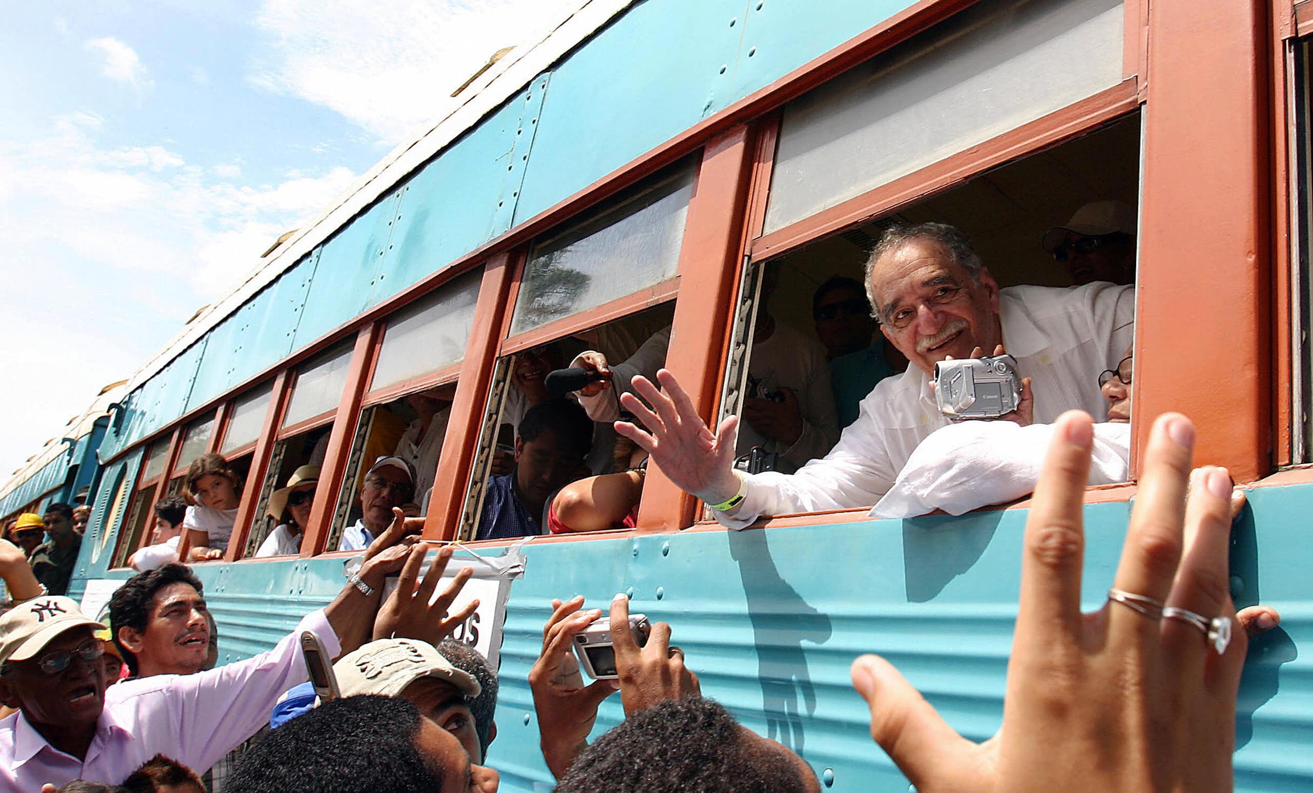 Older man with mustache waves out of a bus window as a crowd gathers.
