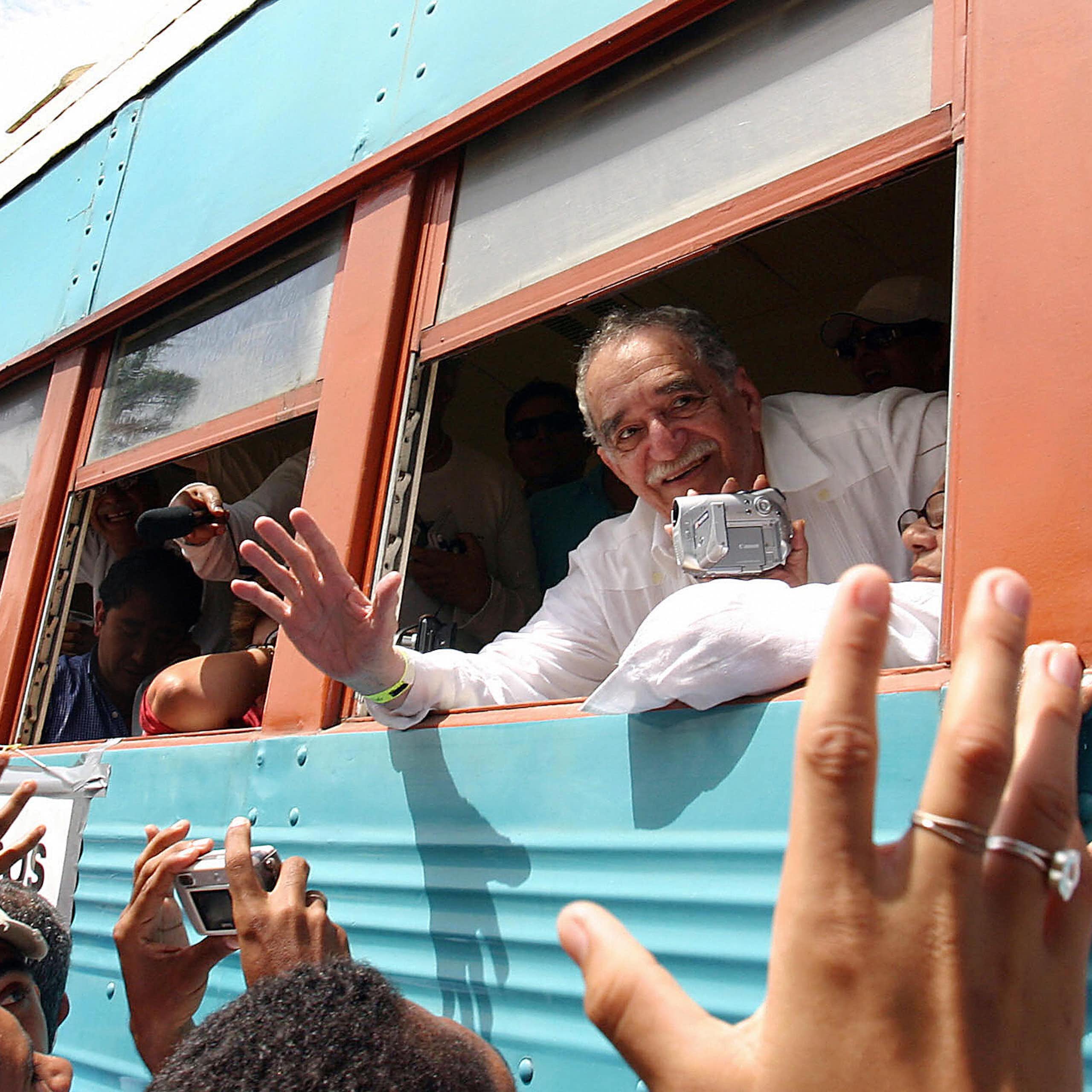 Older man with mustache waves out of a bus window as a crowd gathers.