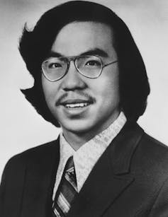 A young Asian man wearing glasses, a jacket and a tie. His hair is fairly long and parted on the side