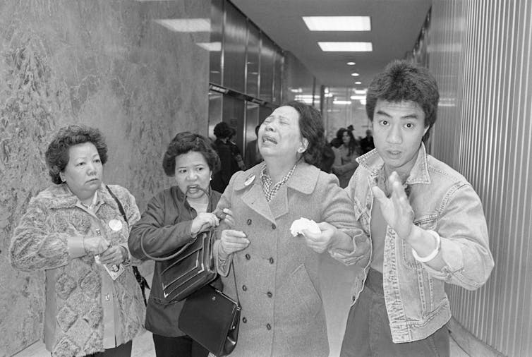 A middle-aged Asian woman throws her head back and wails. Two woman and a young man with spiked hair stand with her and support her.