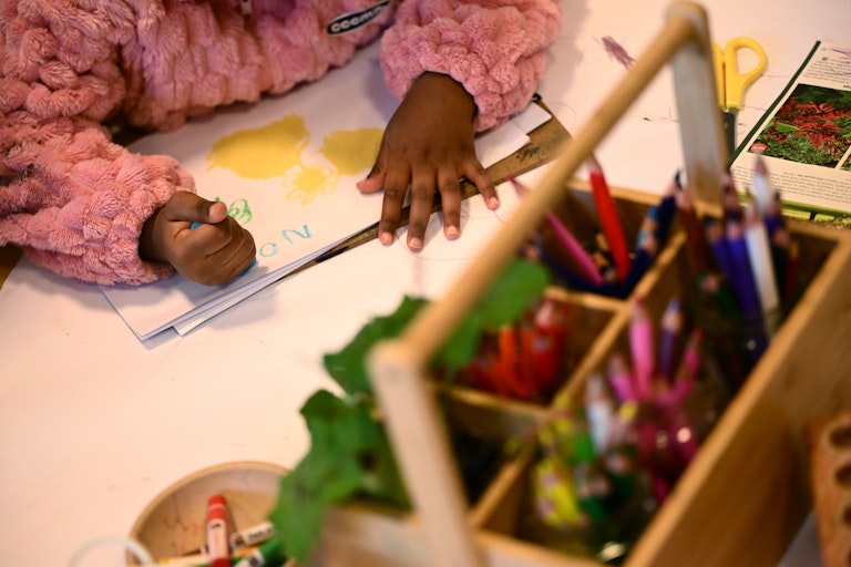 A child draws at a table with a box of pencils nearby.