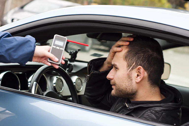 Un homme est assis dans une voiture, la main sur le front, tandis qu'une autre personne lui tend un éthylotest par la fenêtre.