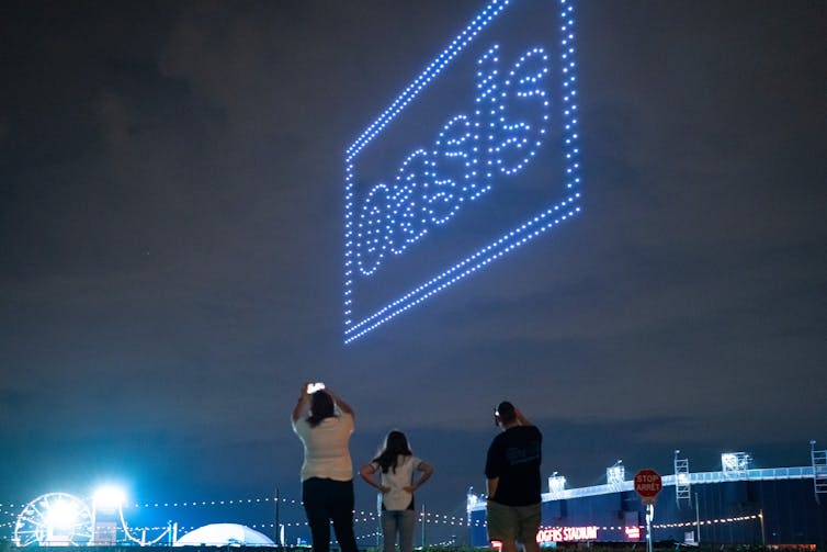 Los drones hacen un logotipo de Oasis en el cielo nocturno mientras la gente pinta fotos desde el suelo.