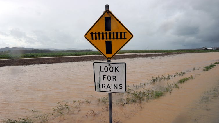 A rail crossing sign reading 'Look for trains' in front of a flooded train line.