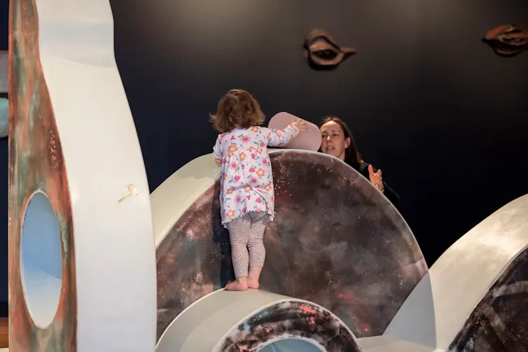 A toddler balances on a curved surface, placing a column while watched over by mum.